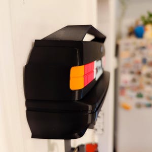 May include: Black key holder shaped like a car's rear end, mounted on a white wall. The key holder features orange, red, and white taillight details. Keys hang from the bottom. A small spoiler is on top.