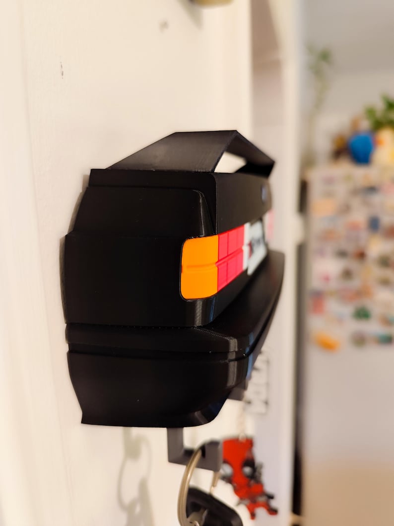 May include: Black key holder shaped like a car's rear end, mounted on a white wall. The key holder features orange, red, and white taillight details. Keys hang from the bottom. A small spoiler is on top.