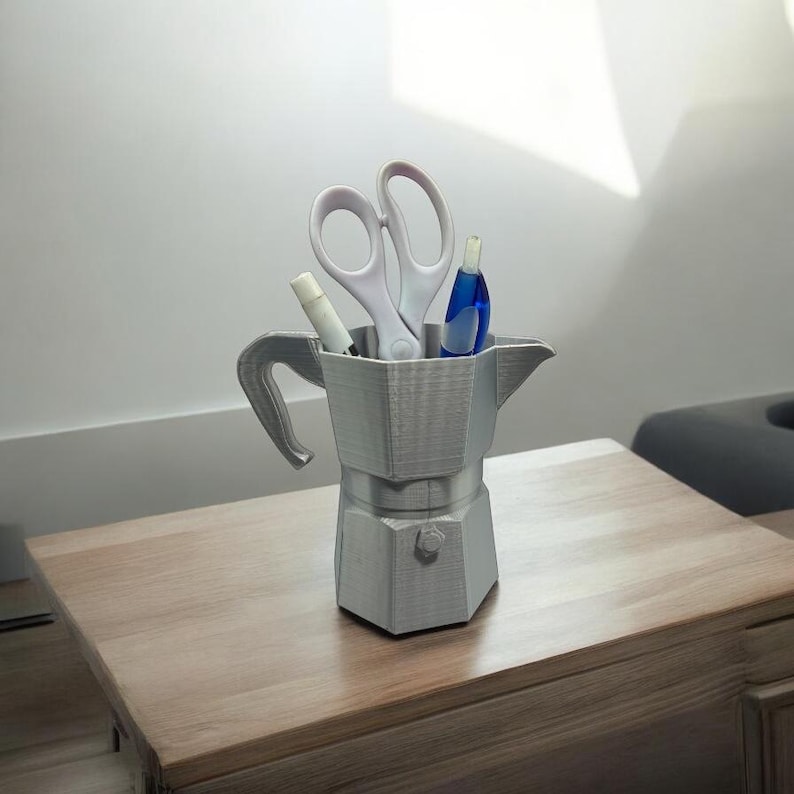 May include: A gray, 3D-printed desk organizer shaped like a stovetop espresso maker. The organizer holds white scissors, a white marker, and a blue pen. The organizer sits on a wooden table.