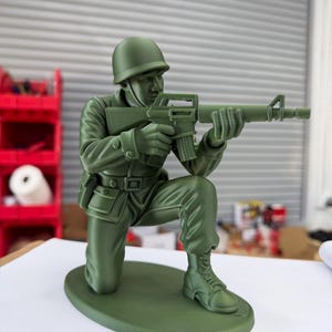 May include: A green plastic toy soldier in a kneeling position, holding a rifle. The soldier wears a helmet, uniform, and boots. The figure is on a green oval base.