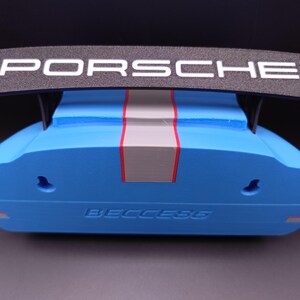 K&ouml;nnte beinhalten: Ein blau-schwarzes Dekorationsobjekt mit dem Wort "PORSCHE" in Wei&szlig; auf einem schwarzen Fl&uuml;gel. Das Objekt hat einen grauen und roten Streifen in der Mitte und das Wort "BECCESG" ist unten sichtbar.