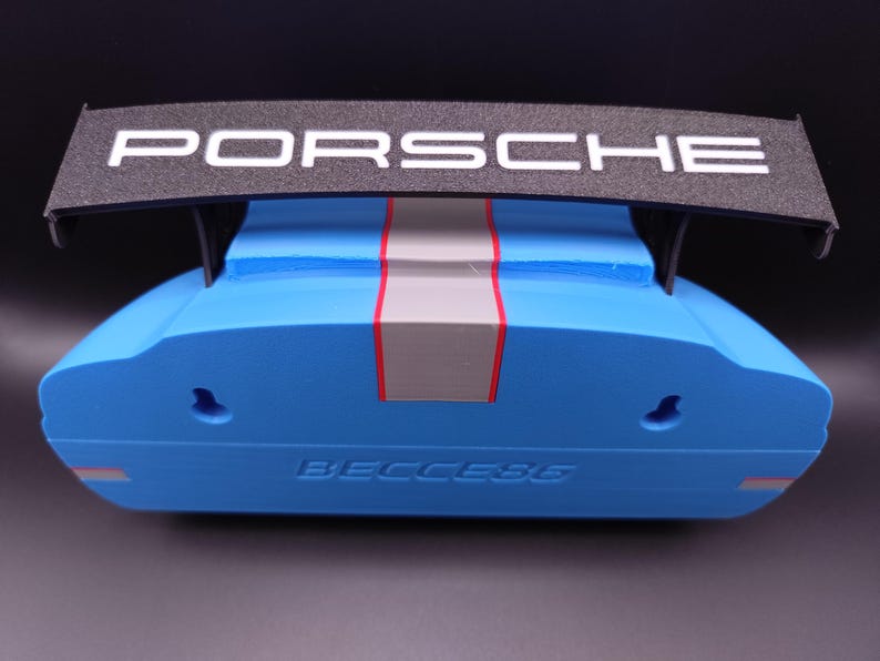 K&ouml;nnte beinhalten: Ein blau-schwarzes Dekorationsobjekt mit dem Wort "PORSCHE" in Wei&szlig; auf einem schwarzen Fl&uuml;gel. Das Objekt hat einen grauen und roten Streifen in der Mitte und das Wort "BECCESG" ist unten sichtbar.