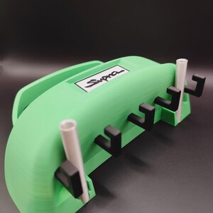 May include: A green 3D-printed organizer with the word "Supra" in white. The organizer has a handle and several black hooks. Two white cylindrical tubes are also present.