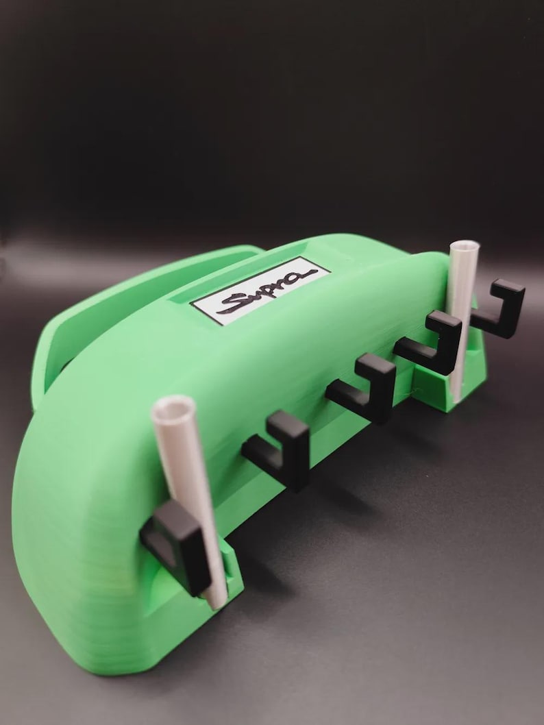 May include: A green 3D-printed organizer with the word "Supra" in white. The organizer has a handle and several black hooks. Two white cylindrical tubes are also present.