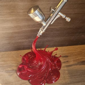 May include: A silver airbrush with a red paint splash design. The airbrush is angled downward, with the red paint forming a dynamic, fluid shape. The base is a detailed, textured splash.