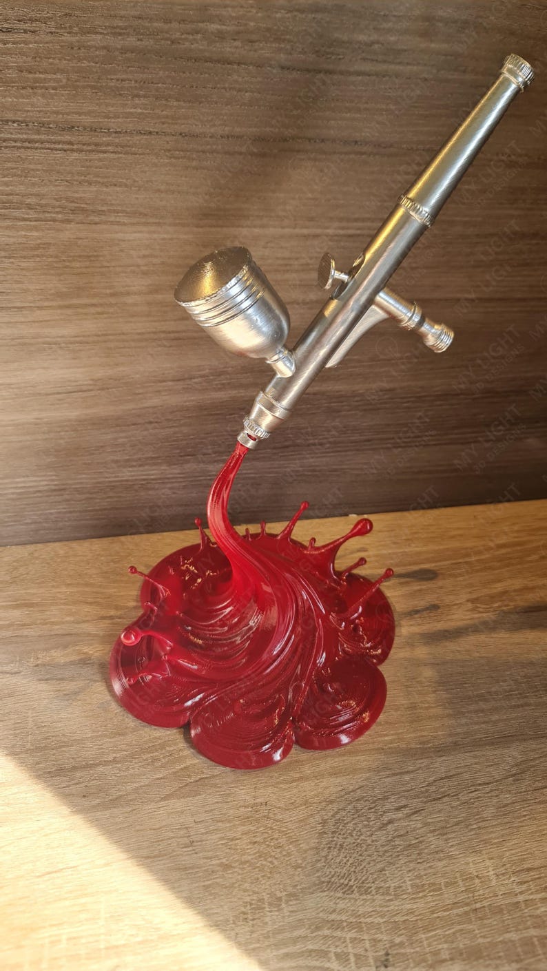 May include: A silver airbrush with a red paint splash design. The airbrush is angled downward, with the red paint forming a dynamic, fluid shape. The base is a detailed, textured splash.