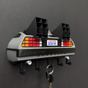 Puede incluir: Un llavero de pared diseñado como la parte trasera de un coche deportivo plateado. Cuenta con dos rejillas de ventilación rectangulares negras, luces traseras rojas y naranjas, y una matrícula blanca que dice CALIFORNIA OUTATIME. Cinco ganchos negros.