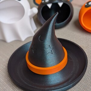 May include: A black witch's hat with an orange band and star and crescent moon details sits on a black tray. Other Halloween-themed containers in the background include a white ghost, a black bat, and an orange pumpkin.