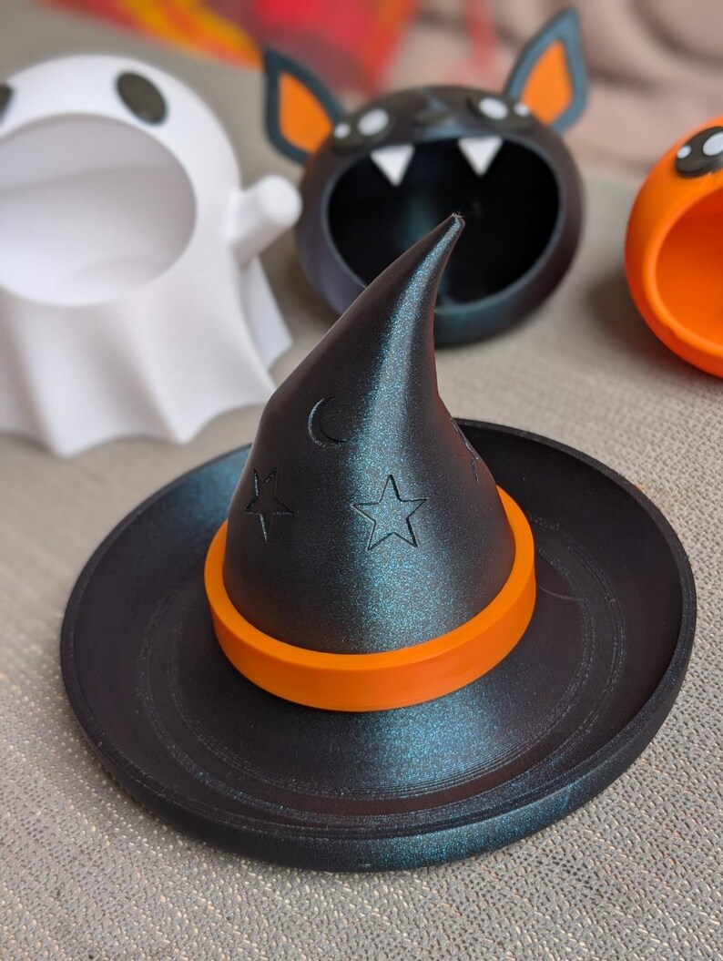 May include: A black witch's hat with an orange band and star and crescent moon details sits on a black tray. Other Halloween-themed containers in the background include a white ghost, a black bat, and an orange pumpkin.