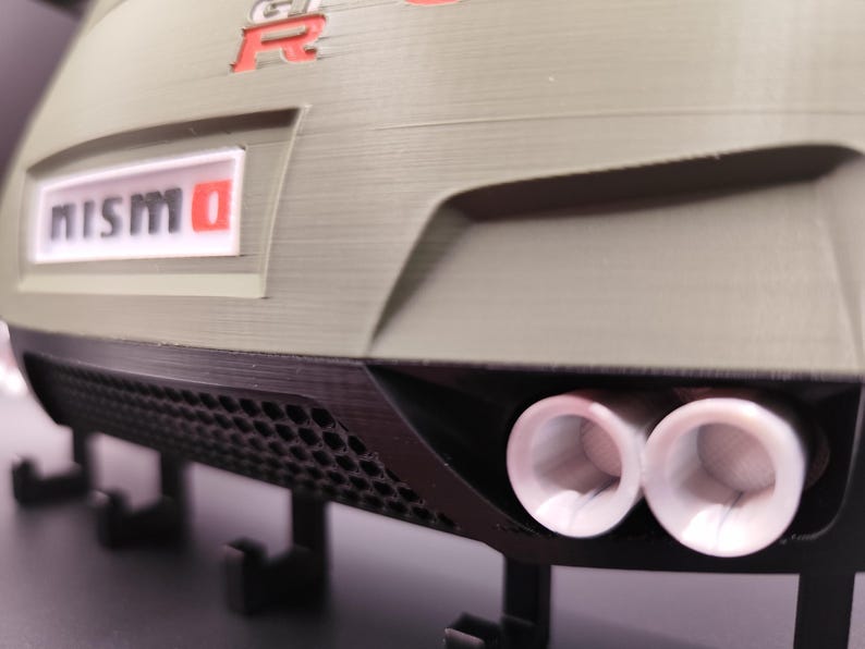 Może przedstawiać: Zbliżenie na tył modelu samochodu z białym i czerwonym logo "NISMO". Samoch&oacute;d jest oliwkowozielony z podw&oacute;jnymi rurami wydechowymi i czarną kratką o strukturze plastra miodu. Emblemat "R" jest czerwony.