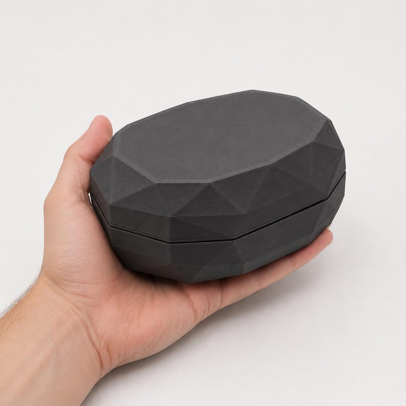 Puede incluir: Una caja negra mate de forma geom&eacute;trica con un dise&ntilde;o facetado. La caja se sostiene en una mano sobre un fondo blanco. La caja parece estar hecha de un material s&oacute;lido y tiene una abertura con bisagras.