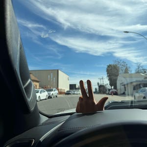 Puede incluir: Un adorno de salpicadero de coche con forma de mano que muestra el signo de la paz. El adorno es de color beige y est&aacute; sobre un salpicadero negro. El fondo muestra un cielo azul con nubes, una calle y coches aparcados.