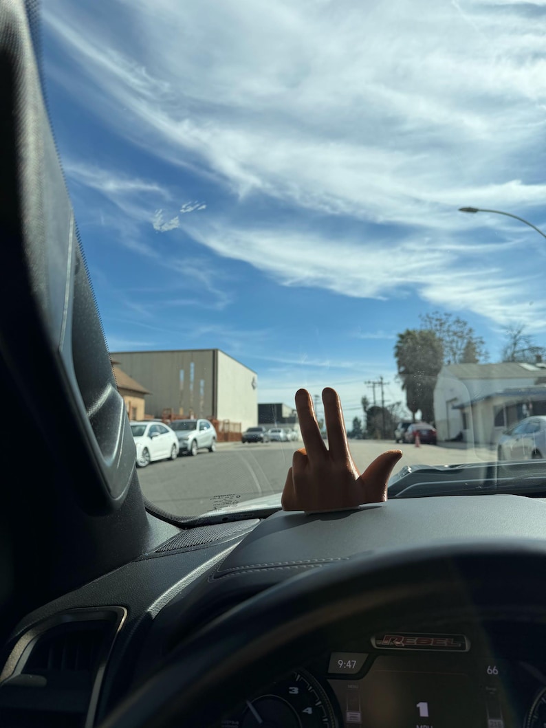Puede incluir: Un adorno de salpicadero de coche con forma de mano que muestra el signo de la paz. El adorno es de color beige y est&aacute; sobre un salpicadero negro. El fondo muestra un cielo azul con nubes, una calle y coches aparcados.