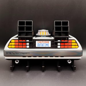 Pode incluir: Um modelo impresso em 3D da m&aacute;quina do tempo DeLorean da s&eacute;rie de filmes De Volta para o Futuro. O modelo apresenta a ic&oacute;nica placa de matr&iacute;cula "OUTATIME", aberturas pretas e uma carro&ccedil;aria prateada. O modelo tem uma base preta com ganchos.