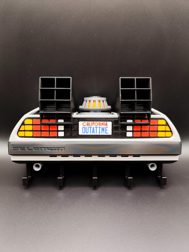 Pode incluir: Um modelo impresso em 3D da m&aacute;quina do tempo DeLorean da s&eacute;rie de filmes De Volta para o Futuro. O modelo apresenta a ic&oacute;nica placa de matr&iacute;cula "OUTATIME", aberturas pretas e uma carro&ccedil;aria prateada. O modelo tem uma base preta com ganchos.