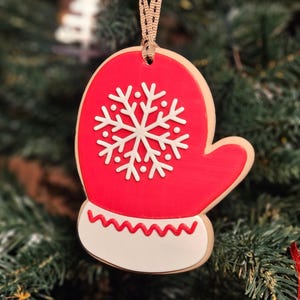 K&ouml;nnte beinhalten: Ein roter, handschuhf&ouml;rmiger Weihnachtsschmuck mit einem wei&szlig;en Schneeflockenmuster. Der Schmuck hat einen wei&szlig;en Boden mit einem roten Zickzackmuster. Er h&auml;ngt an einem beigen Band vor einem verschwommenen Weihnachtsbaumhintergrund.