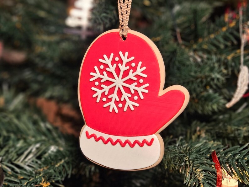 K&ouml;nnte beinhalten: Ein roter, handschuhf&ouml;rmiger Weihnachtsschmuck mit einem wei&szlig;en Schneeflockenmuster. Der Schmuck hat einen wei&szlig;en Boden mit einem roten Zickzackmuster. Er h&auml;ngt an einem beigen Band vor einem verschwommenen Weihnachtsbaumhintergrund.