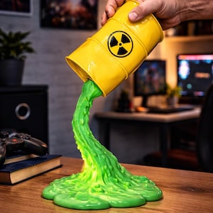 May include: A yellow barrel, marked with a black radiation symbol, is depicted pouring out a stream of bright green, viscous liquid. The liquid flows onto a wooden surface, forming a puddle. The barrel is held by a hand.