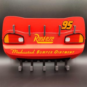 May include: Red wall-mounted key holder shaped like Lightning McQueen's rear end, with the number 95 and the text "Rust-eze Medicated Bumper Ointment". It has five gray hooks for keys.