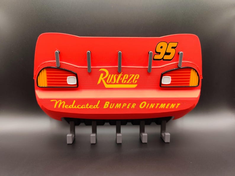 May include: Red wall-mounted key holder shaped like Lightning McQueen's rear end, with the number 95 and the text "Rust-eze Medicated Bumper Ointment". It has five gray hooks for keys.