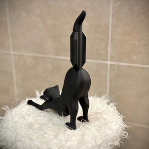 Pode incluir: Estatueta de gato preto em pose brincalhona, com as costas arqueadas e a cauda levantada. O gato est&aacute; sobre uma superf&iacute;cie branca e fofa. A cauda tem um design geom&eacute;trico &uacute;nico.
