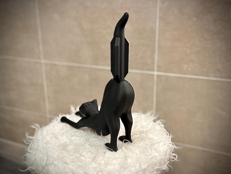 Pode incluir: Estatueta de gato preto em pose brincalhona, com as costas arqueadas e a cauda levantada. O gato est&aacute; sobre uma superf&iacute;cie branca e fofa. A cauda tem um design geom&eacute;trico &uacute;nico.