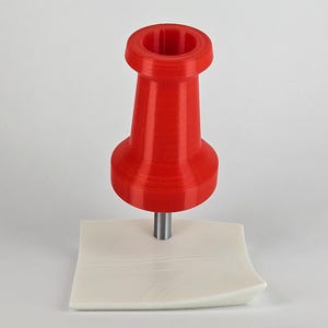 May include: A bright red push pin with a cylindrical body and a flat head, mounted on a small, off-white, square base. The pin is made of plastic and has a metallic pin. The background is plain white.