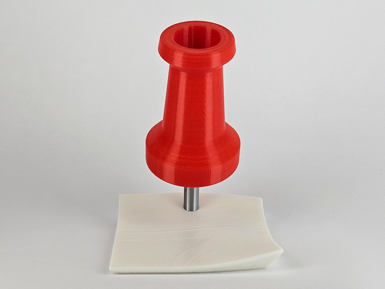 May include: A bright red push pin with a cylindrical body and a flat head, mounted on a small, off-white, square base. The pin is made of plastic and has a metallic pin. The background is plain white.