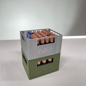 May include: Two stacked battery storage containers, one silver and one olive green, each labeled with "AAA" and "AA" respectively. The silver container holds numerous AAA batteries, while the green container holds AA batteries. The batteries are copper and blue.
