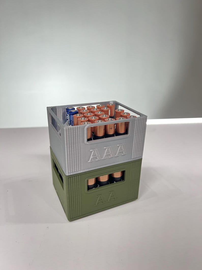 May include: Two stacked battery storage containers, one silver and one olive green, each labeled with "AAA" and "AA" respectively. The silver container holds numerous AAA batteries, while the green container holds AA batteries. The batteries are copper and blue.