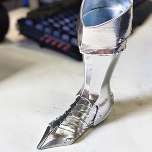 May include: A silver-coloured, pointed-toe boot with a high cuff. The boot has a metallic sheen and is designed with layered panels and rivets. The interior of the cuff is a lighter shade of silver.