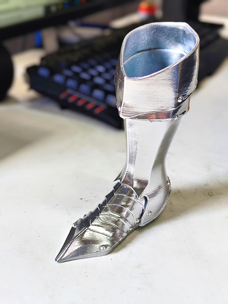 May include: A silver-coloured, pointed-toe boot with a high cuff. The boot has a metallic sheen and is designed with layered panels and rivets. The interior of the cuff is a lighter shade of silver.