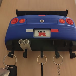 May include: A blue car-shaped key holder with a black spoiler and exhaust pipes. The key holder has a license plate that reads "GT R". The key holder has four hooks for keys and keychains.