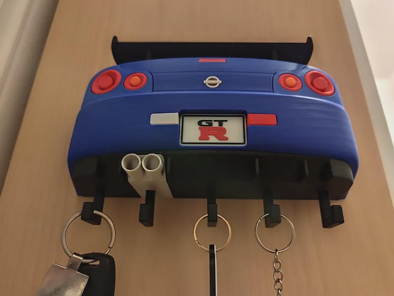 May include: A blue car-shaped key holder with a black spoiler and exhaust pipes. The key holder has a license plate that reads "GT R". The key holder has four hooks for keys and keychains.