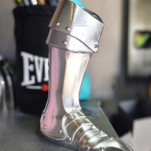 May include: A silver-coloured, metallic leg armour piece. The armour has a pointed toe and is detailed with rivets and layered plates. The leg armour is designed to protect the lower leg and ankle. The background includes a black container with the word "EVERLAST" on it.