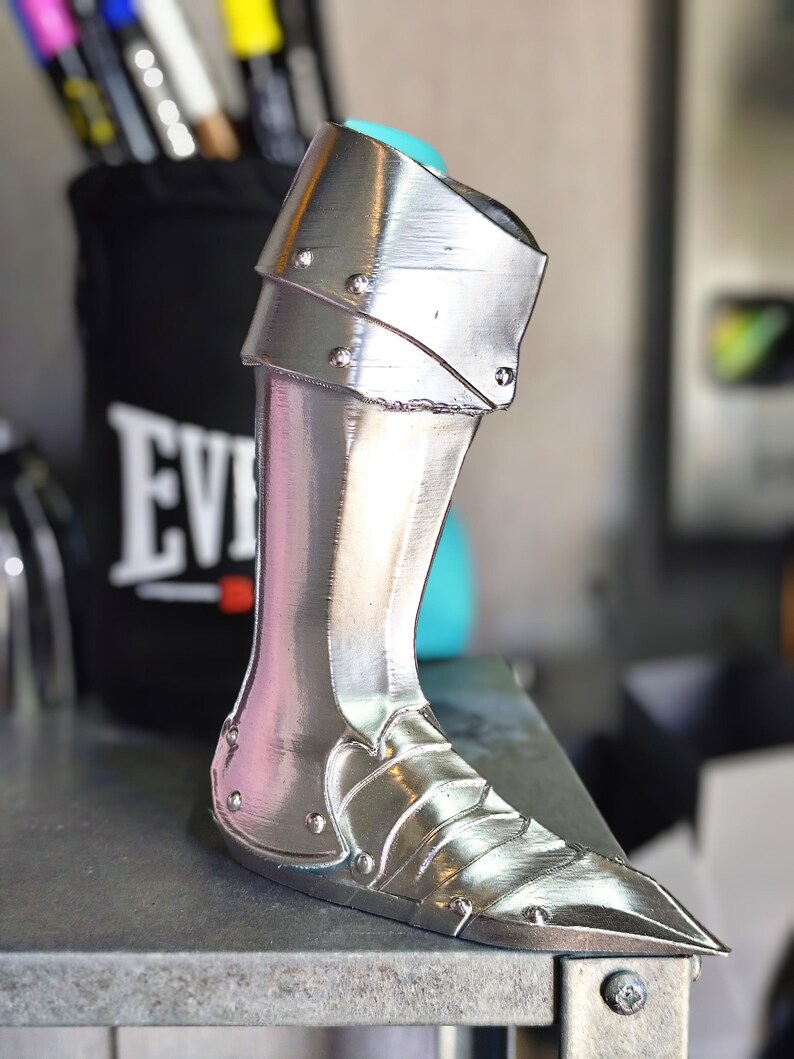 May include: A silver-coloured, metallic leg armour piece. The armour has a pointed toe and is detailed with rivets and layered plates. The leg armour is designed to protect the lower leg and ankle. The background includes a black container with the word "EVERLAST" on it.