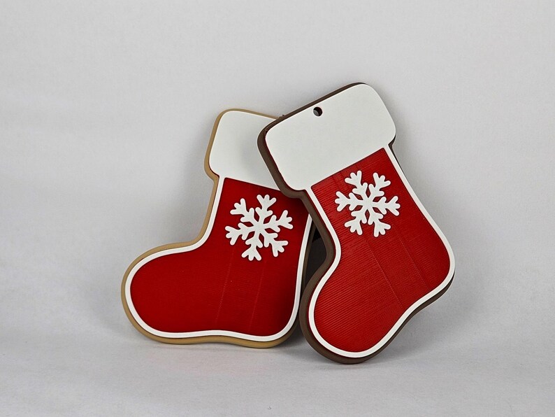 K&ouml;nnte beinhalten: Zwei Weihnachtsstrumpf-Ornamente. Die Str&uuml;mpfe sind rot mit wei&szlig;en Oberteilen und einem wei&szlig;en Schneeflocken-Design. Einer hat einen braunen Rand, der andere einen beigen Rand. Beide haben ein Loch zum Aufh&auml;ngen.