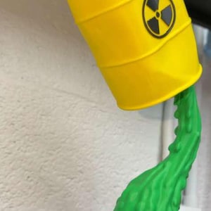 May include: A yellow barrel with a black and yellow radiation symbol, tilted to pour out green liquid. The barrel is a decorative item, possibly a lamp or sculpture, with a unique design.