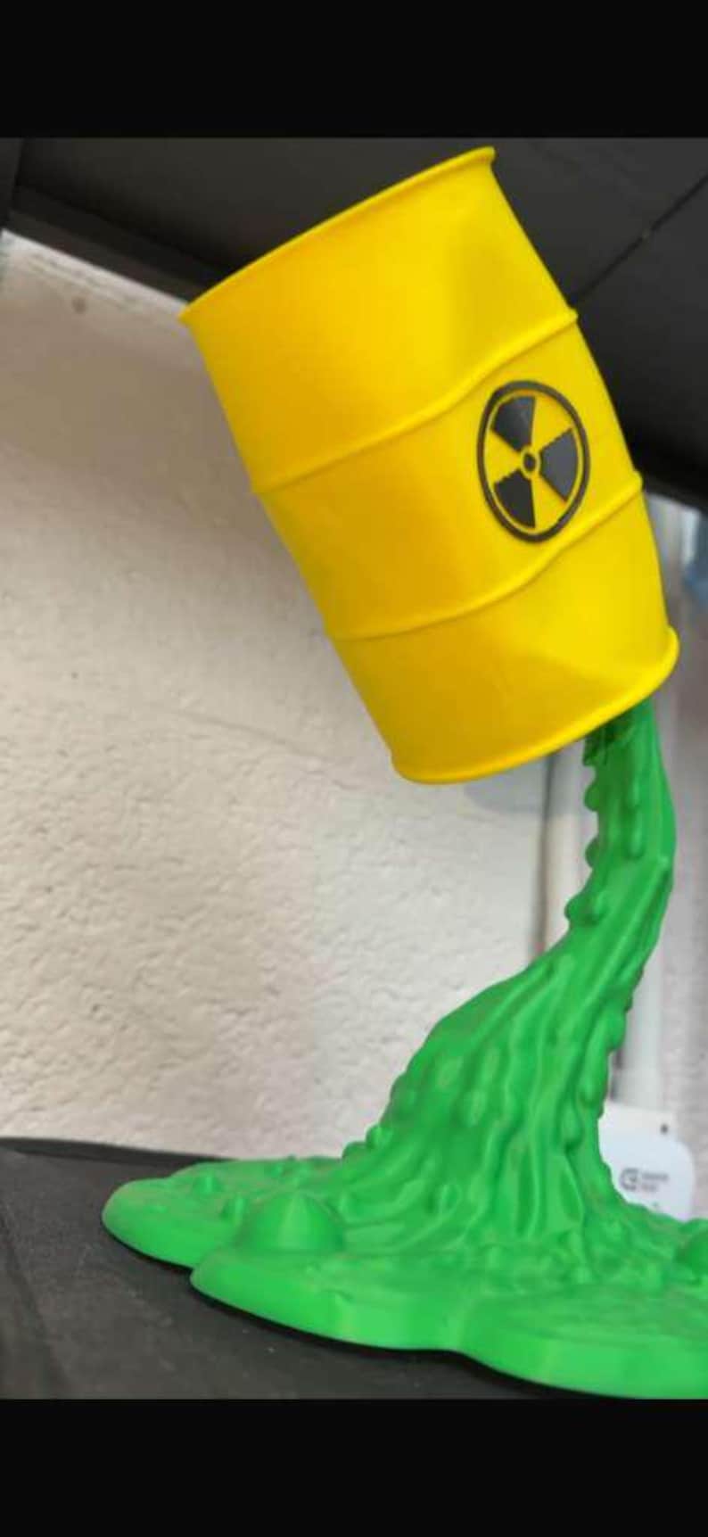 May include: A yellow barrel with a black and yellow radiation symbol, tilted to pour out green liquid. The barrel is a decorative item, possibly a lamp or sculpture, with a unique design.