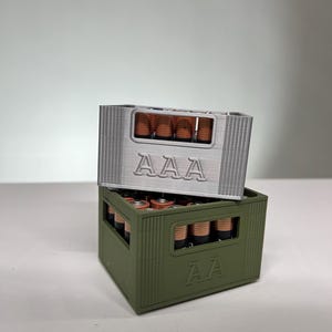 May include: Two 3D-printed battery holders, one silver and one olive green, stacked. Each holder is designed to resemble a crate and is labelled with "AAA" on the front. The holders are filled with AAA batteries.