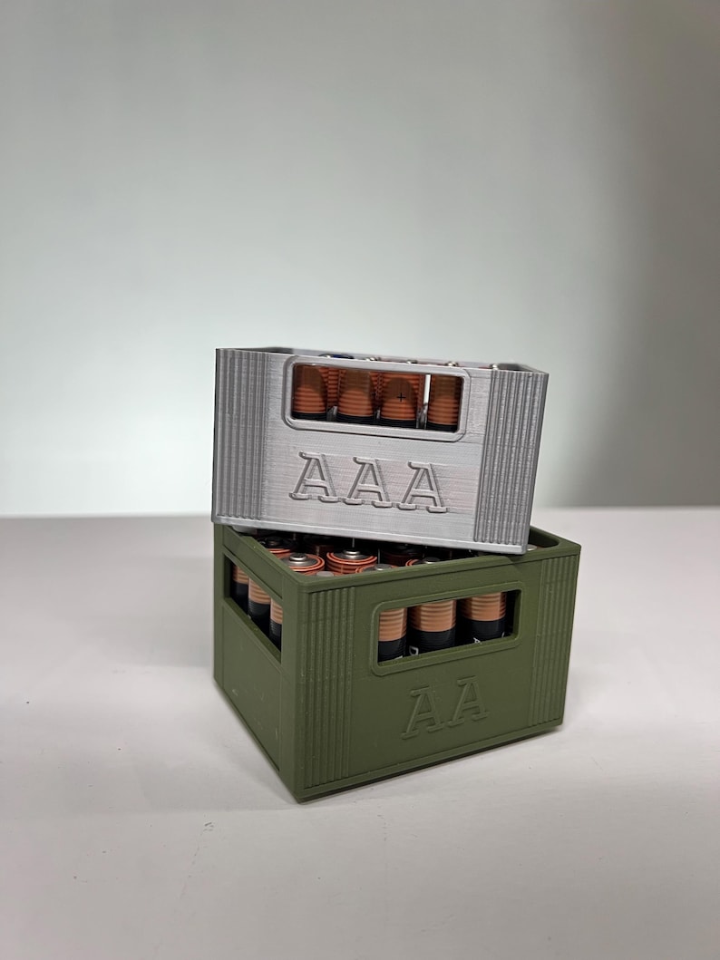 May include: Two 3D-printed battery holders, one silver and one olive green, stacked. Each holder is designed to resemble a crate and is labelled with "AAA" on the front. The holders are filled with AAA batteries.