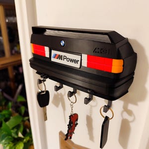 May include: Black key holder shaped like a car's rear end, featuring red, white, and orange taillights. It has the "M Power" logo and a BMW emblem. Includes key hooks with attached keys and keychains.