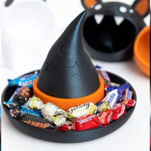 May include: A black witch hat candy dish with star and crescent moon cutouts, filled with wrapped sweets. The hat sits on an orange base and a black plate. Other Halloween-themed containers are in the background.