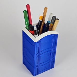 May include: A blue and white plastic pencil holder shaped like a portable toilet. The holder has a white rim and is filled with various pens and pencils.