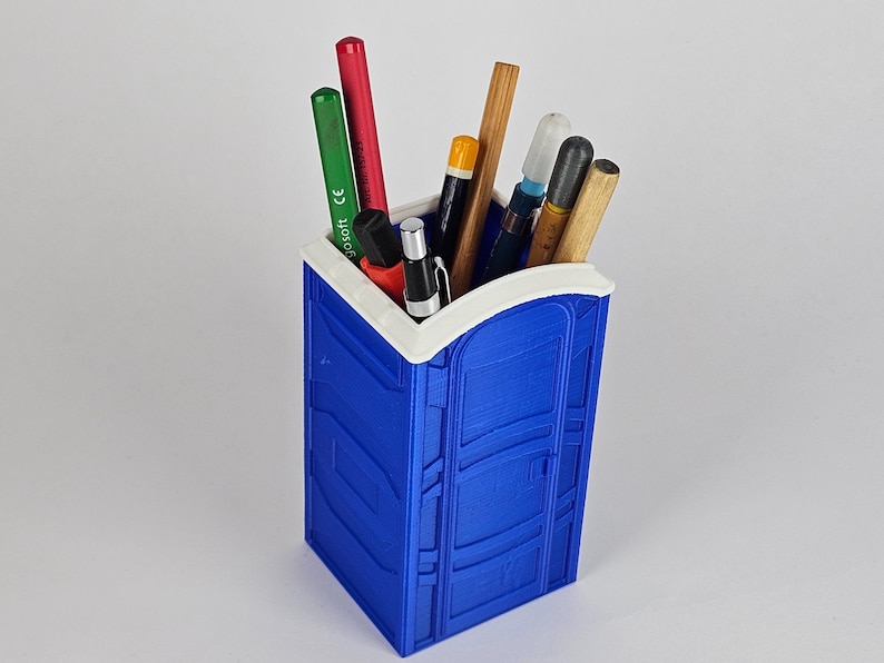 May include: A blue and white plastic pencil holder shaped like a portable toilet. The holder has a white rim and is filled with various pens and pencils.