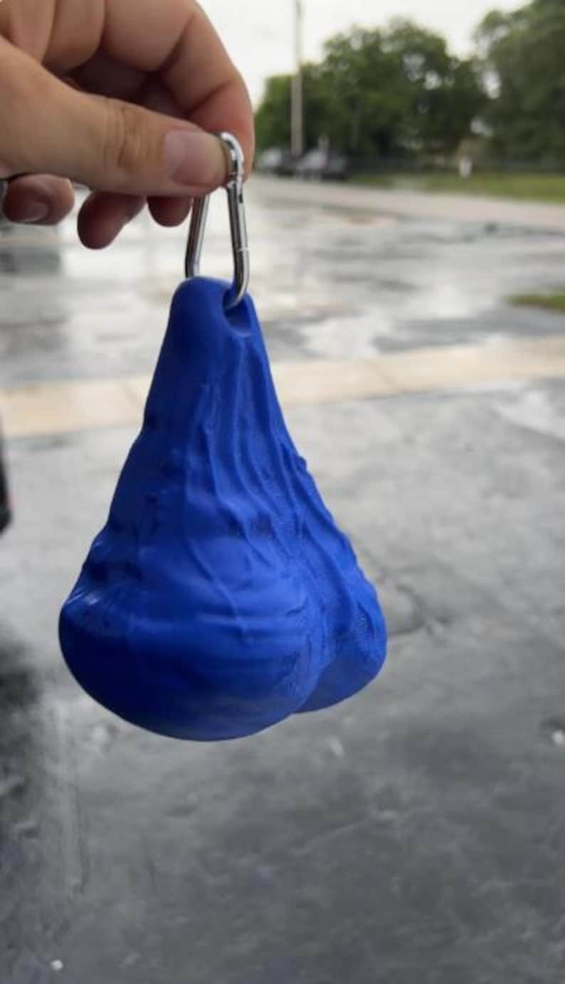 May include: A bright blue, textured, three-dimensional object with a rounded, dual-lobed shape and a wrinkled surface. The object features a small hole at the top, through which a silver-colored metal carabiner clip is attached for hanging.