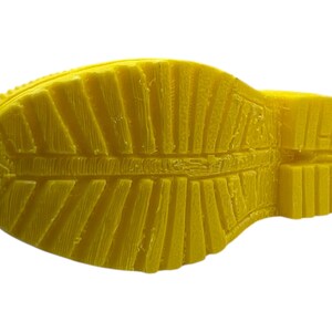 May include: A close-up of the sole of a yellow boot. The sole has a textured pattern with ridges and grooves for traction. The boot is made of a solid, bright yellow material.