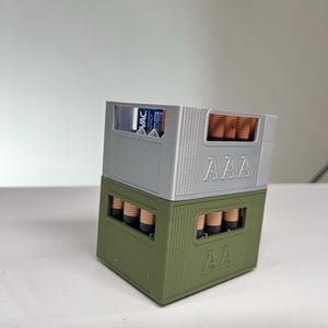 May include: Two 3D-printed battery holders, one silver and one olive green, stacked on top of each other. The top holder is labelled "AAA" and contains AAA batteries. The bottom holder is labelled "AA" and contains AA batteries. The top holder also has a "VAC Energy" label.