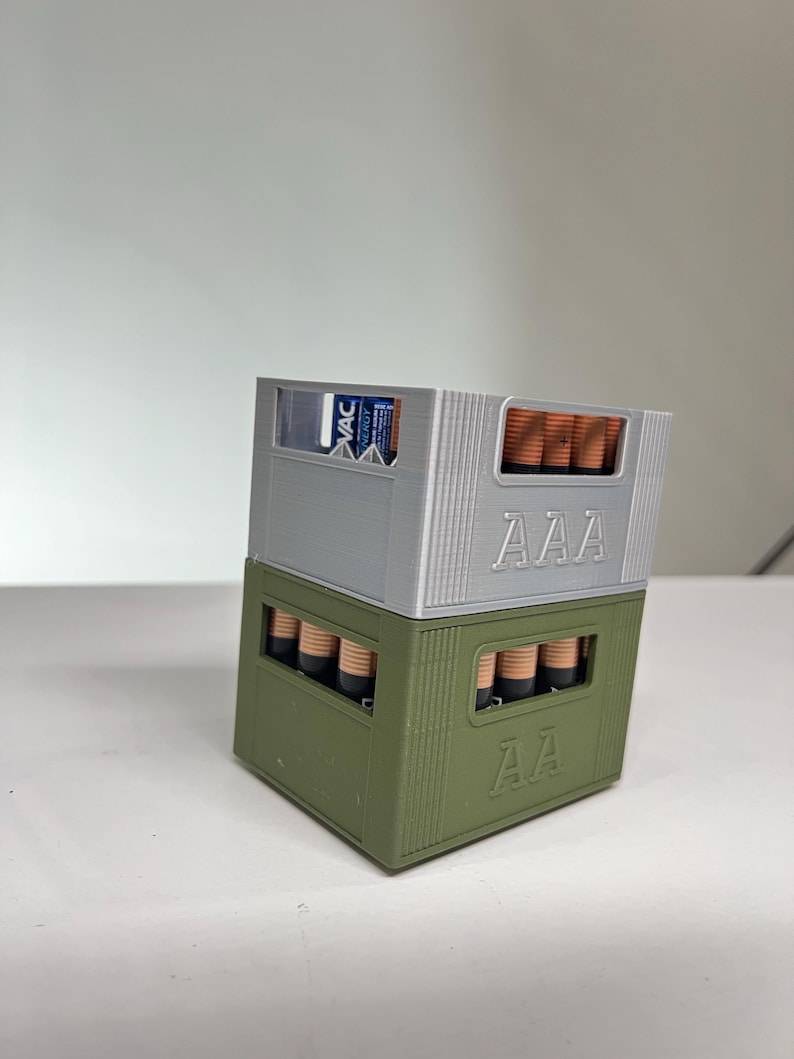 May include: Two 3D-printed battery holders, one silver and one olive green, stacked on top of each other. The top holder is labelled "AAA" and contains AAA batteries. The bottom holder is labelled "AA" and contains AA batteries. The top holder also has a "VAC Energy" label.