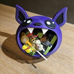 May include: A purple bat-shaped candy bowl filled with various wrapped candies and a lollipop. The bowl has large white fangs, black eyes, and ears. The candies are in colorful wrappers. The bowl sits on a wooden surface.